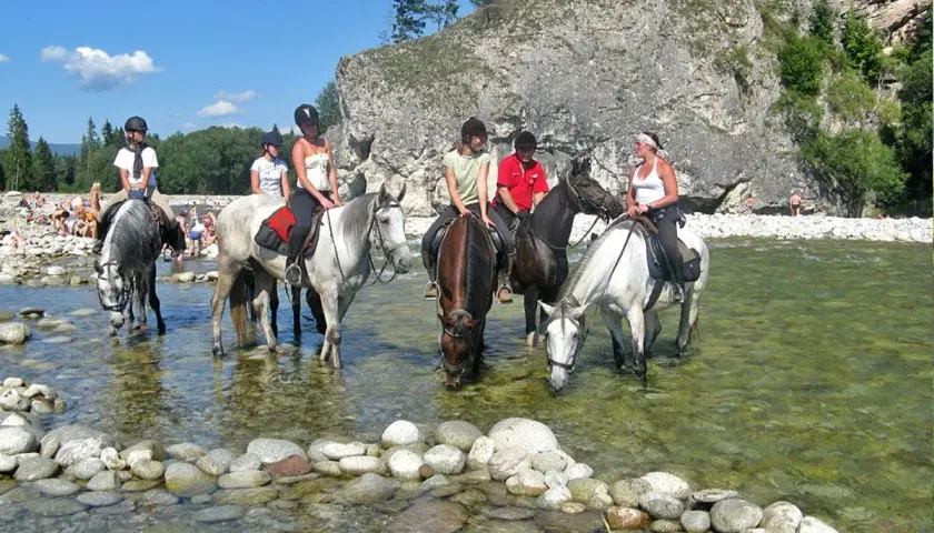 Konni jeźdźcy na koniach w rzece, w tle skaliste zbocze i plaża z ludźmi. To widok z Bukowiny Tatrzańskiej, jakby rynek atrakcji.