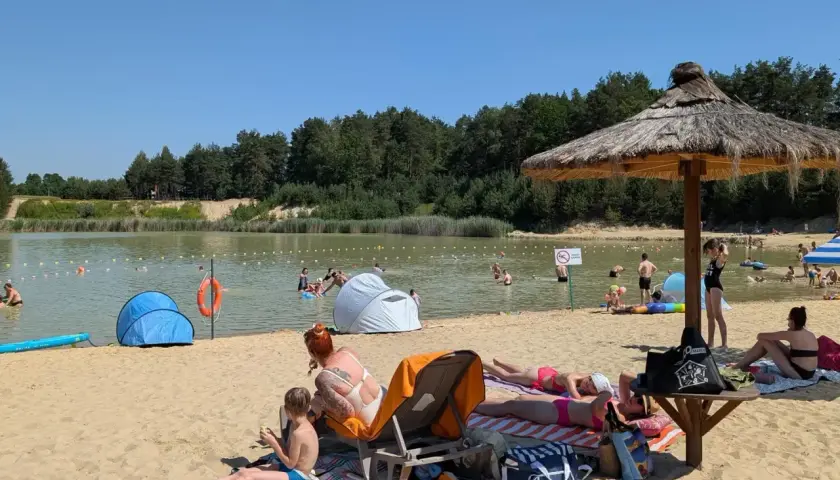 Słoneczny dzień nad zalewem Biszcza. Ludzie opalają się na plaży, kąpią w wodzie i odpoczywają pod parasolami.
