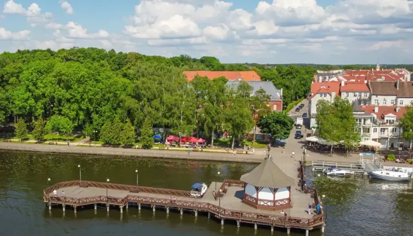 Malownicze miejscowości na Mazurach: drewniane molo z altaną, zielony park i urokliwe kamienice nad jeziorem.