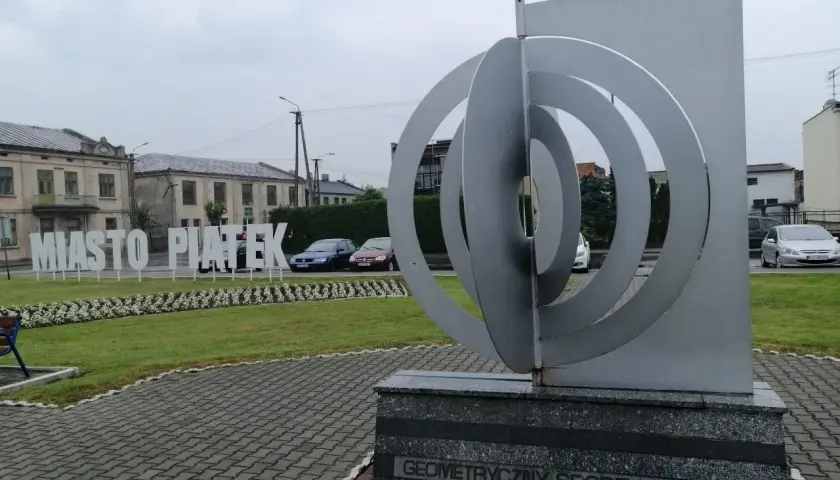 Geometryczny środek Polski w miejscowości Piątek. Monumentalna rzeźba i napis "Miasto Piątek" na tle budynków.