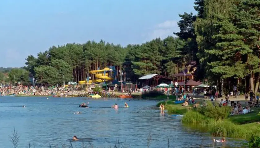 Tłum ludzi wypoczywa nad wodą, widać zjeżdżalnie wodne i drzewa. Krasnobród zalew tętni życiem w słoneczny dzień.