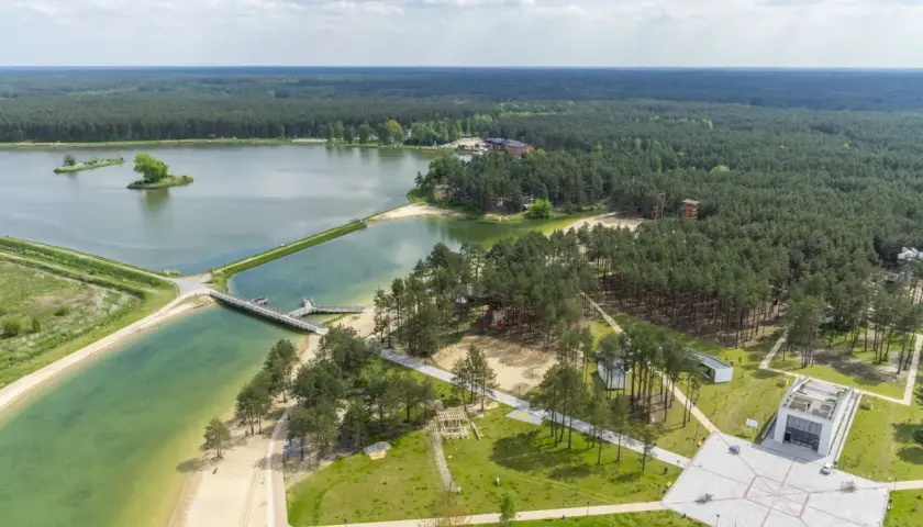 Malowniczy krajobraz z lotu ptaka: rozległy las otacza turkusowe wody zalewu janowskiego, z piaszczystymi plażami i nowoczesną architekturą.