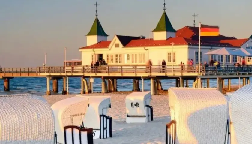Zachód słońca nad molo w Ahlbecku. Białe kosze plażowe na piasku, a w tle budynek z restauracją i niemiecką flagą. To jedne z wielu atrakcji Ahlbecku.