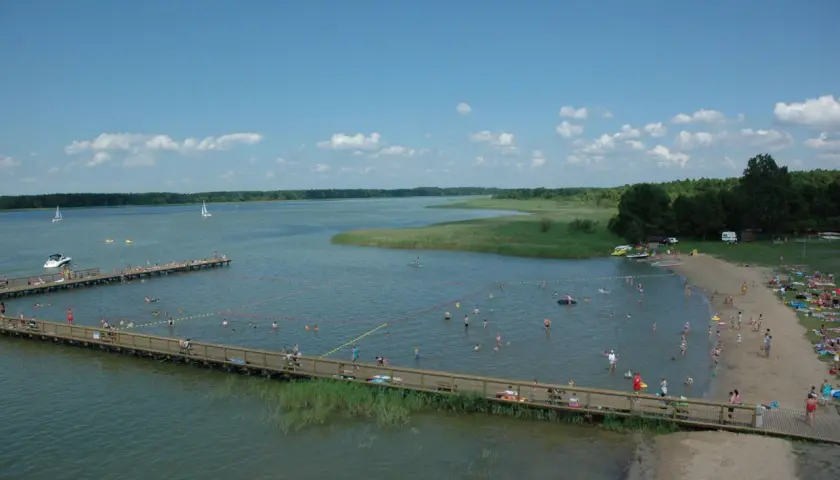 Wypływa z jeziora roś, a plażowicze kąpią się i opalają. W oddali widać żaglówki i łodzie.
