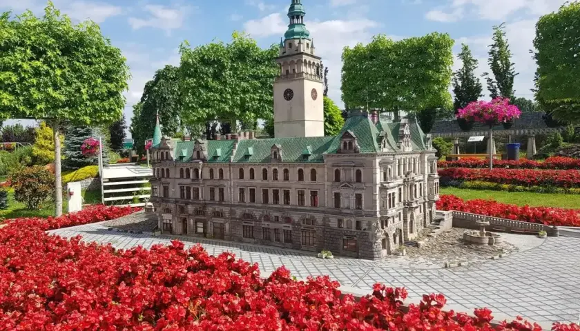 Miniatura ratusza z wieżą zegarową, otoczona czerwonymi kwiatami, w miniaturowej Kotlinie Kłodzkiej.