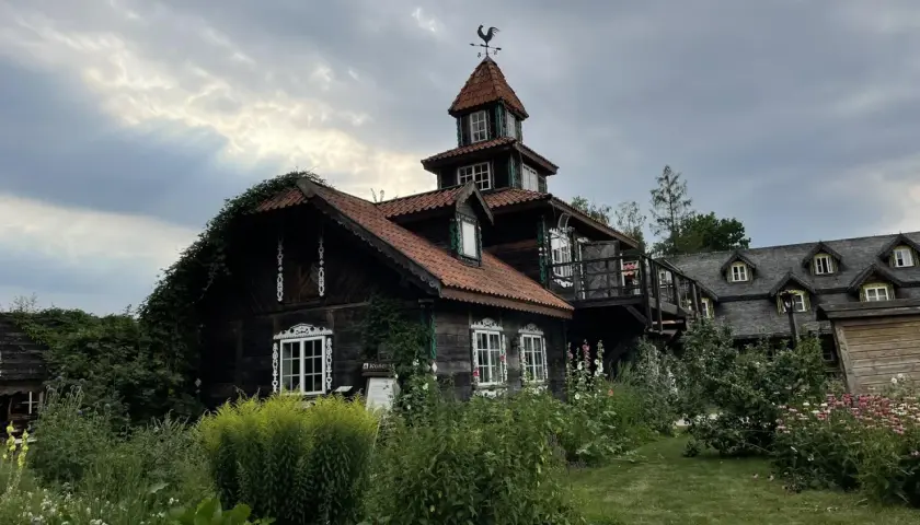 Drewniany dom z wieżyczką i kogutem na dachu, otoczony bujną zielenią. W oddali widać drugi budynek z dormerami.