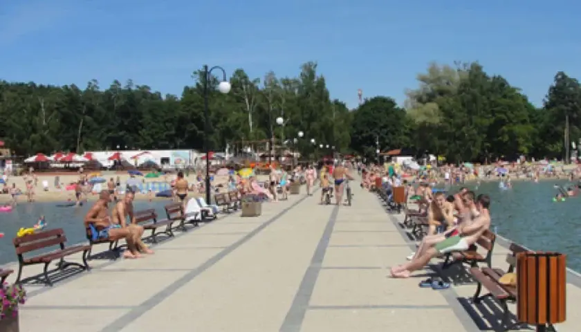 Ludzie odpoczywają na pomoście nad Skorzęcinem, jezioro tętni życiem, plaża pełna plażowiczów.