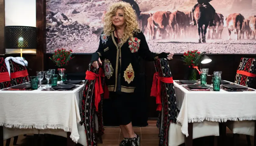 Kobieta w ozdobnej marynarce stoi przy stole nakrytym do kolacji. W tle obraz z kowbojem i bydłem. To zapowiedź mammy food w Łodzi.