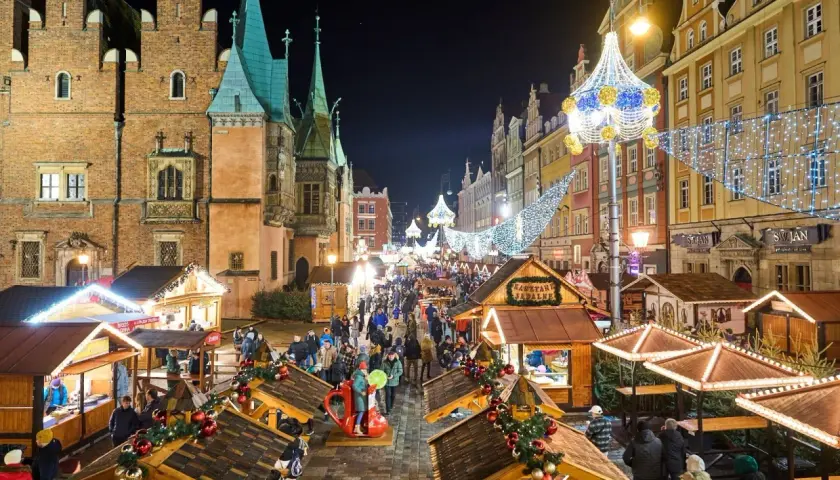 Tłoczny jarmark we Wrocławiu nocą, udekorowany świątecznymi światłami. Drewniane stragany oferują przysmaki, a tłumy spacerują wśród kolorowych kamienic.