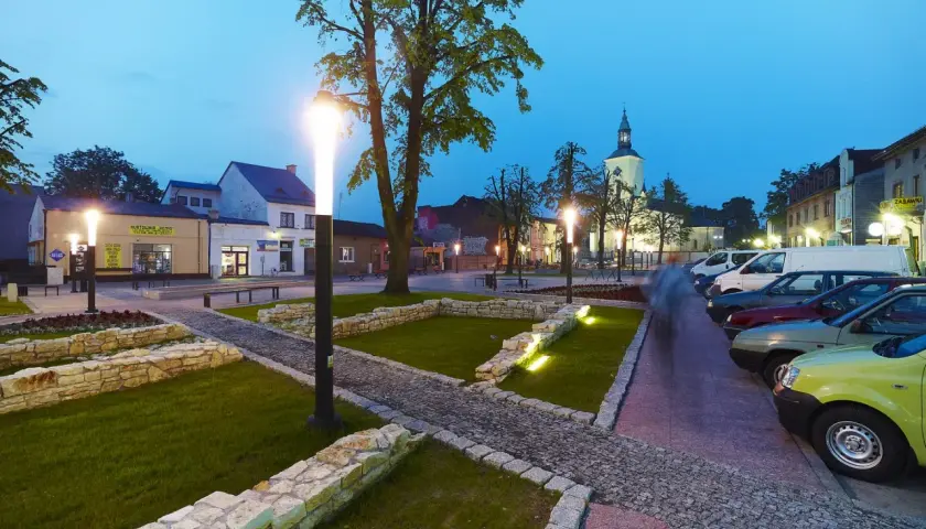 Rynek w Żarkach Letnisko o zmierzchu, z oświetlonymi latarniami, brukowanymi alejkami i zaparkowanymi samochodami.