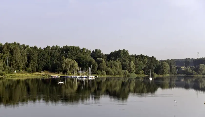 Zalew Przeczyce z łodziami przy brzegu i samotną białą żaglówką na spokojnej tafli wody.