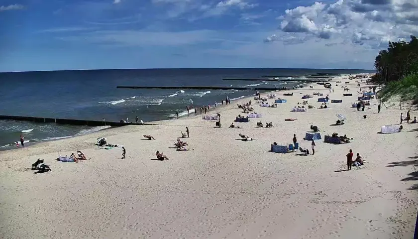 Słoneczny dzień na plaży w Dźwirzynie. Ludzie odpoczywają na piasku, kąpią się w morzu i spacerują po deptaku.