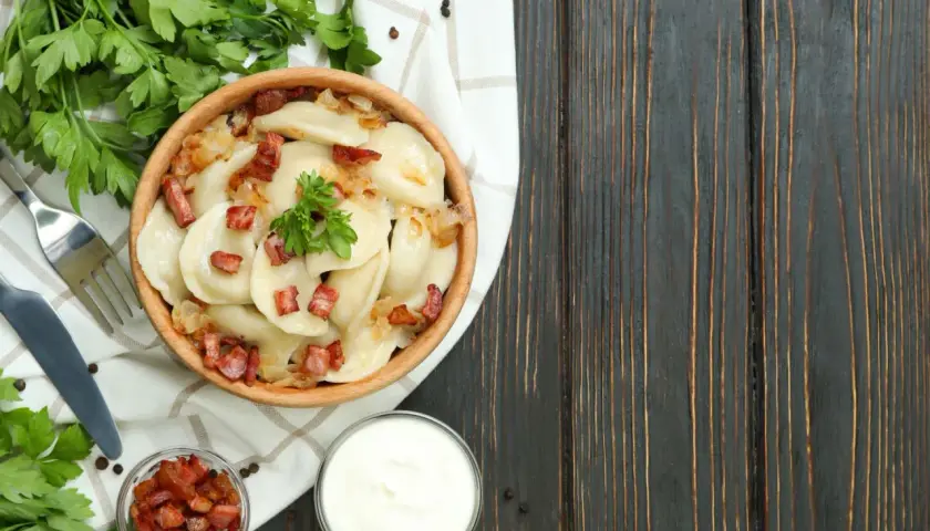 Pyszne pierogi z boczkiem i cebulką, serwowane w drewnianej misie. To kwintesencja kuchni łemkowskiej, podane z kwaśną śmietaną i świeżą natką pietruszki.