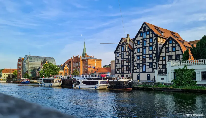 Bydgoszcz wydarzenia dzisiaj: na Wyspie Młyńskiej spaceruje po linie figura, cumują statki, a w tle widać zabytkowe kamienice i nowoczesną architekturę.