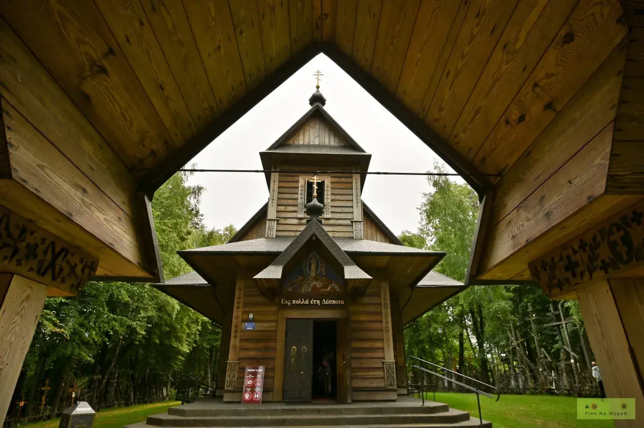 Drewniana cerkiew z krzyżami na tle zieleni. W oddali widać wiele krzyży, tworzących niezwykłą grabarkę.