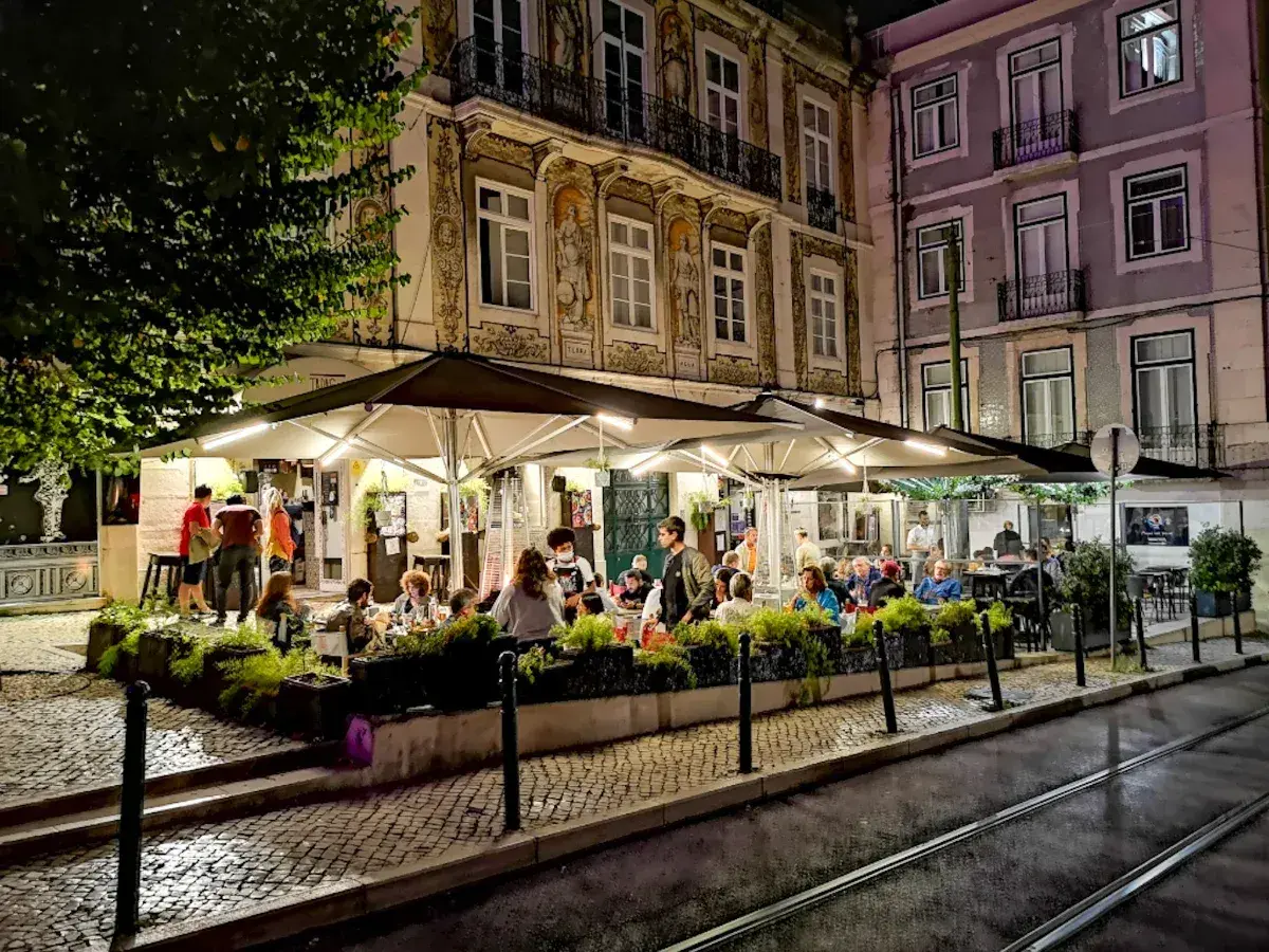 Wieczorny gwar w **Bairro Alto**. Ludzie siedzą przy stolikach pod parasolami, delektując się posiłkiem i atmosferą.