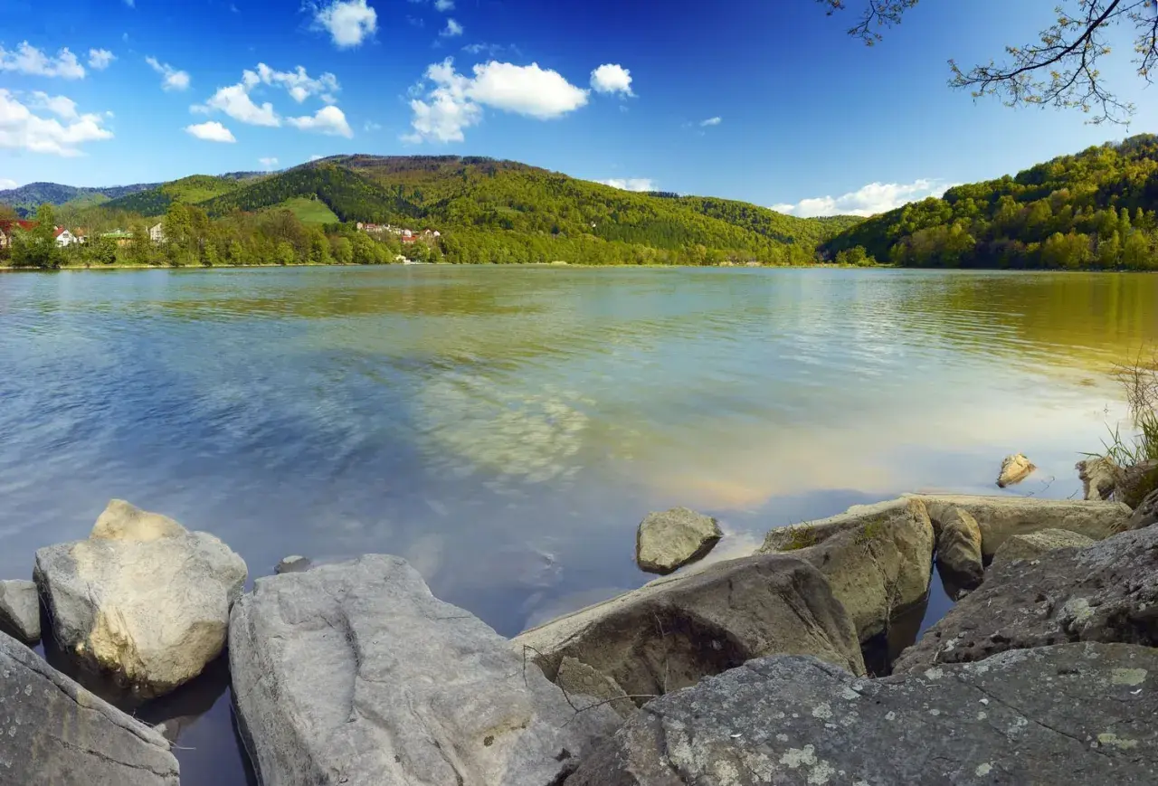 Kamienista plaża nad Jeziorem Międzybrodzkim, otoczona zielonymi wzgórzami i błękitnym niebem z białymi chmurami.