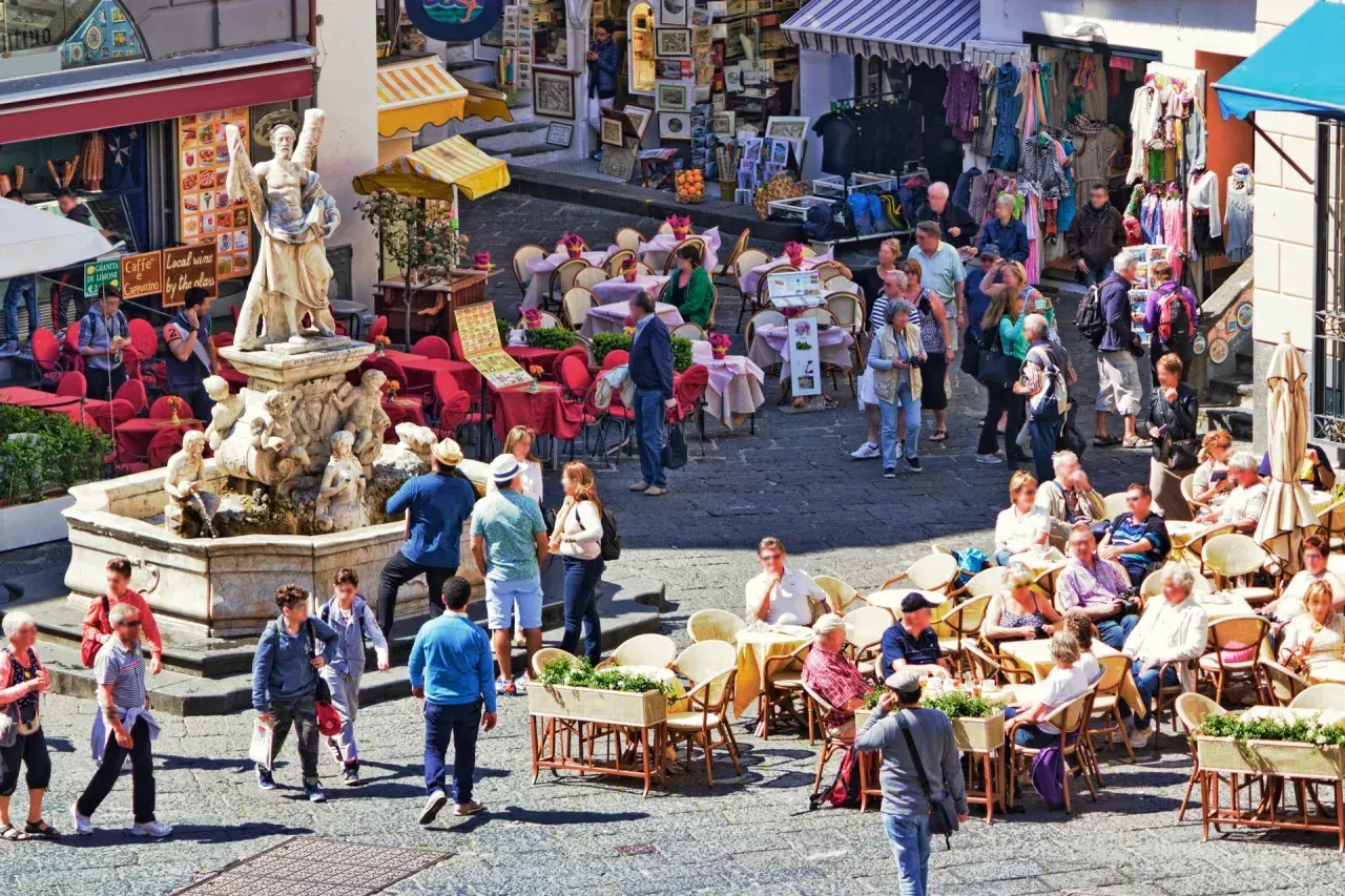 Tłoczny plac z fontanną i rzeźbą, kawiarnie z czerwonymi stolikami i ludźmi siedzącymi na zewnątrz, sklep z ubraniami w tle. Wszystko pod słońcem, tworząc wakacyjny, **coperto** nastrój.