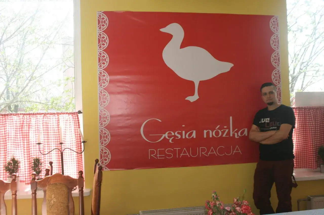 Mężczyzna stoi obok wielkiego czerwonego banera z białym gęsią i napisem "Gęsia nóżka RESTAURACJA".