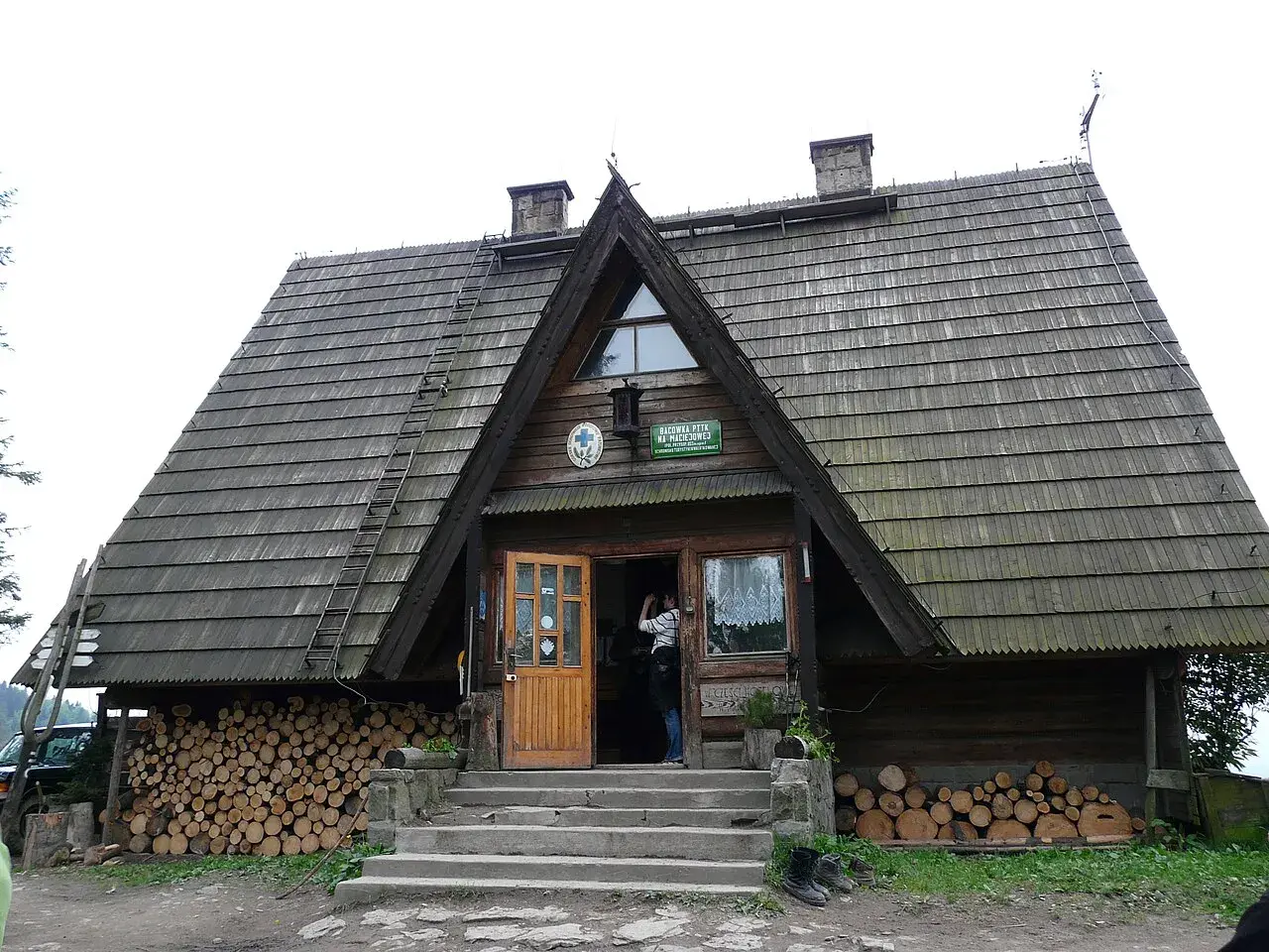 Drewniane schronisko Maciejowa z otwartymi drzwiami, stosy drewna po bokach i drabina na dachu.