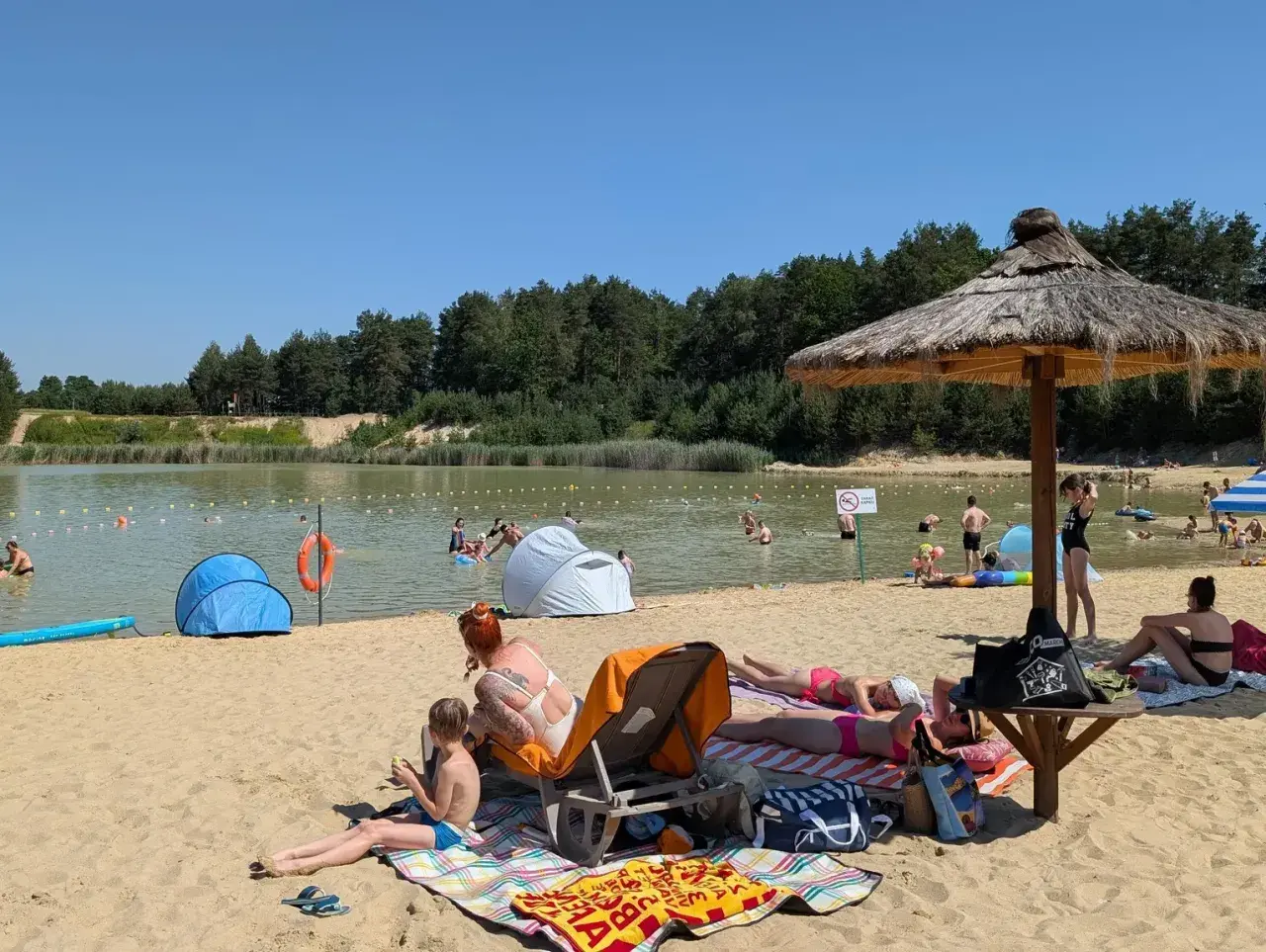 Słoneczny dzień nad zalewem Biszcza. Ludzie opalają się na plaży, kąpią w wodzie i odpoczywają pod parasolami.