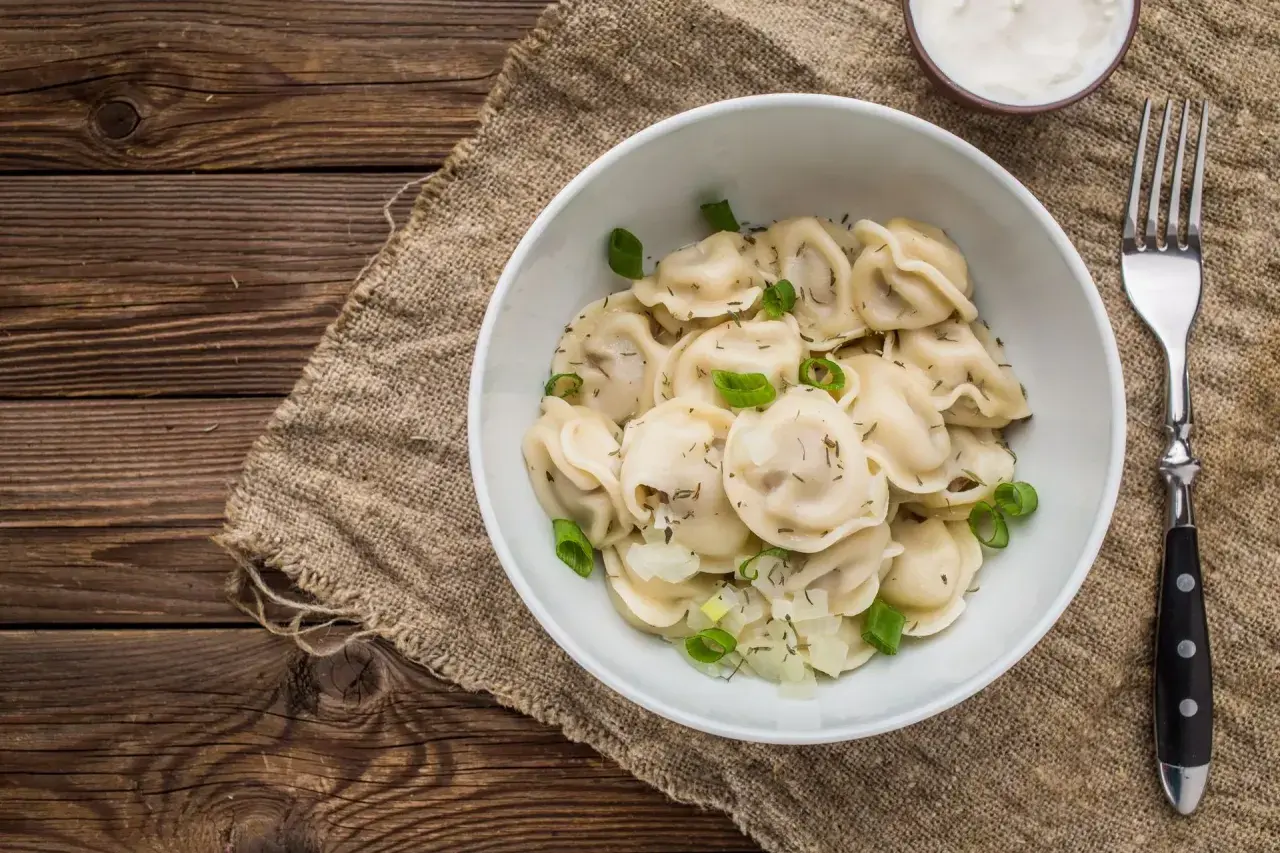 Pyszne pierogi z cebulką i koperkiem, idealne na obiad w stylu zapiecek warszawa. Obok miseczka śmietany i widelec.