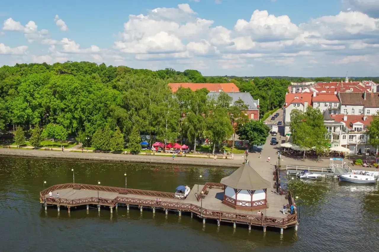 Malownicze miejscowości na Mazurach: drewniane molo z altaną, zielony park i urokliwe kamienice nad jeziorem.