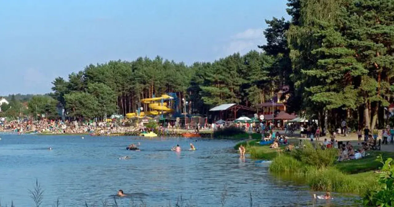 Tłum ludzi wypoczywa nad wodą, widać zjeżdżalnie wodne i drzewa. Krasnobród zalew tętni życiem w słoneczny dzień.