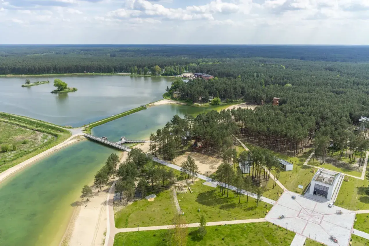 Malowniczy krajobraz z lotu ptaka: rozległy las otacza turkusowe wody zalewu janowskiego, z piaszczystymi plażami i nowoczesną architekturą.
