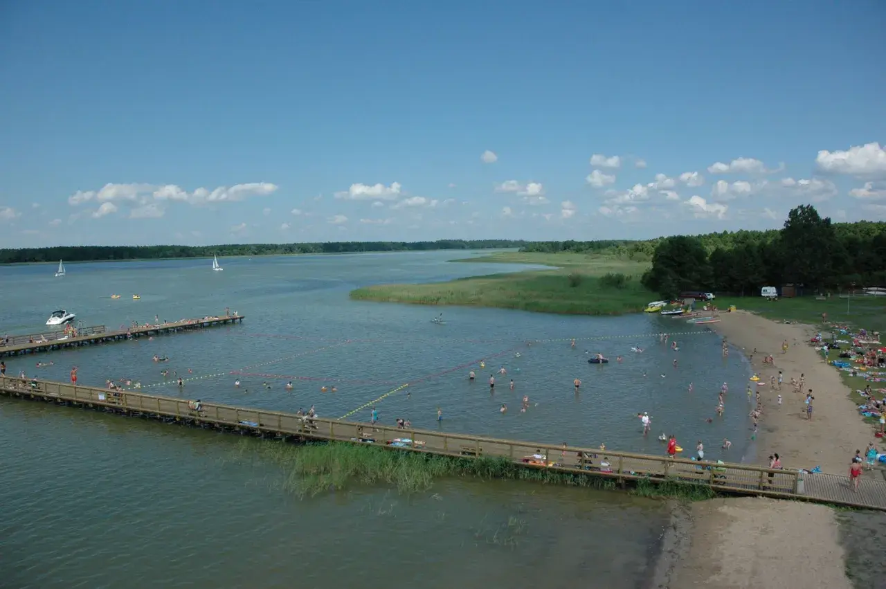 Wypływa z jeziora roś, a plażowicze kąpią się i opalają. W oddali widać żaglówki i łodzie.