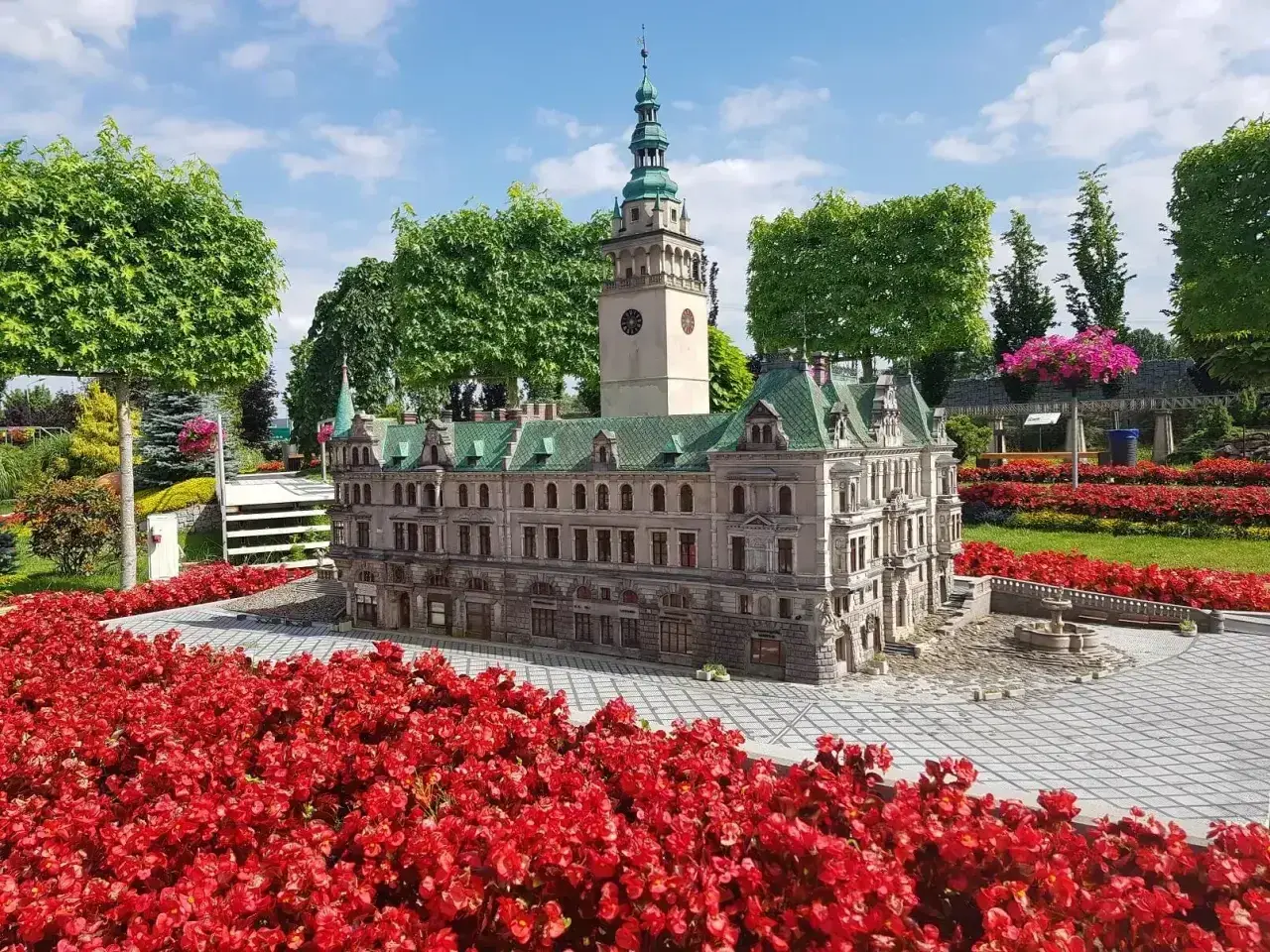 Miniatura ratusza z wieżą zegarową, otoczona czerwonymi kwiatami, w miniaturowej Kotlinie Kłodzkiej.