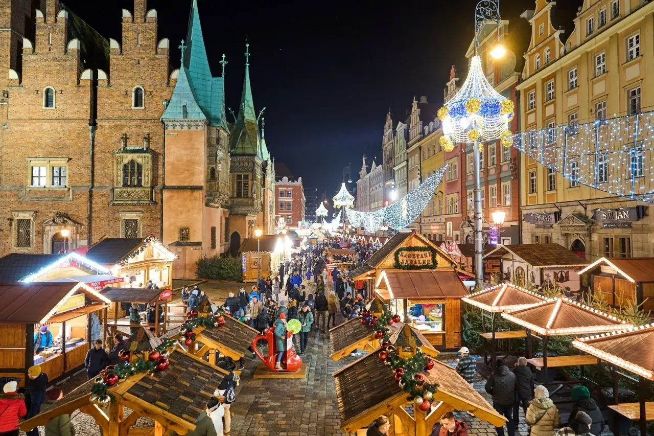 Tłoczny jarmark we Wrocławiu nocą, udekorowany świątecznymi światłami. Drewniane stragany oferują przysmaki, a tłumy spacerują wśród kolorowych kamienic.