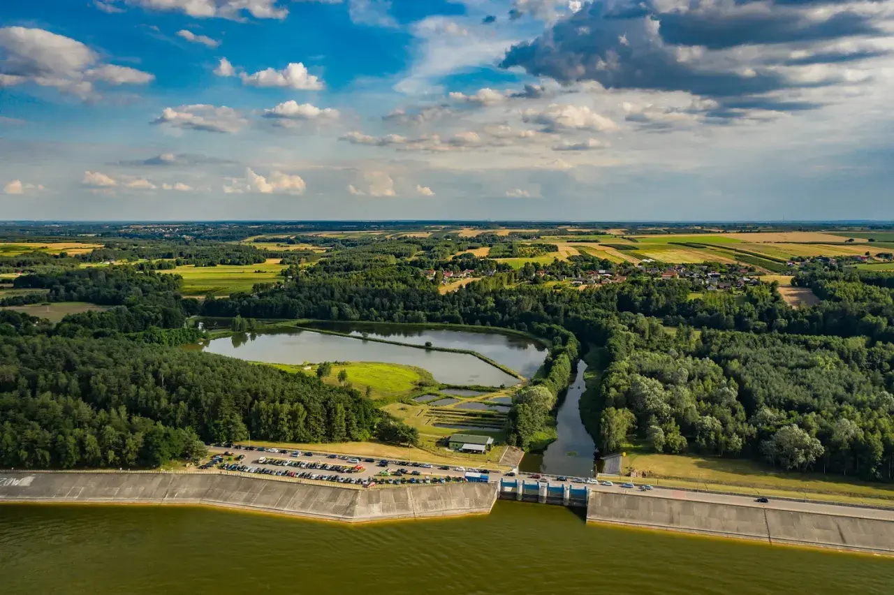 Malowniczy krajobraz z tamą i zbiornikiem wodnym. Jezioro Chańcza otoczone lasami i polami, z widoczną drogą na koronie zapory.