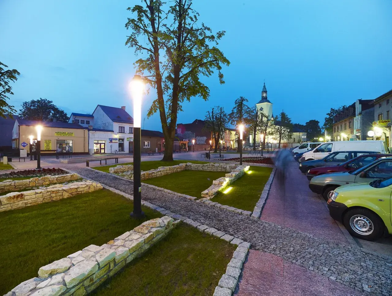 Rynek w Żarkach Letnisko o zmierzchu, z oświetlonymi latarniami, brukowanymi alejkami i zaparkowanymi samochodami.