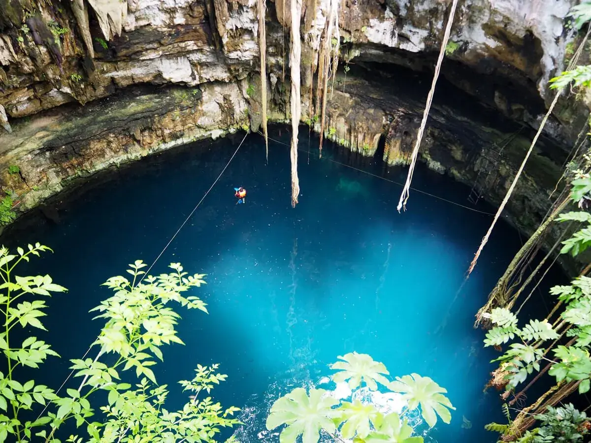 Niesamowity cenote z turkusową wodą, wiszącymi korzeniami i małą postacią pływającą w głębinach.