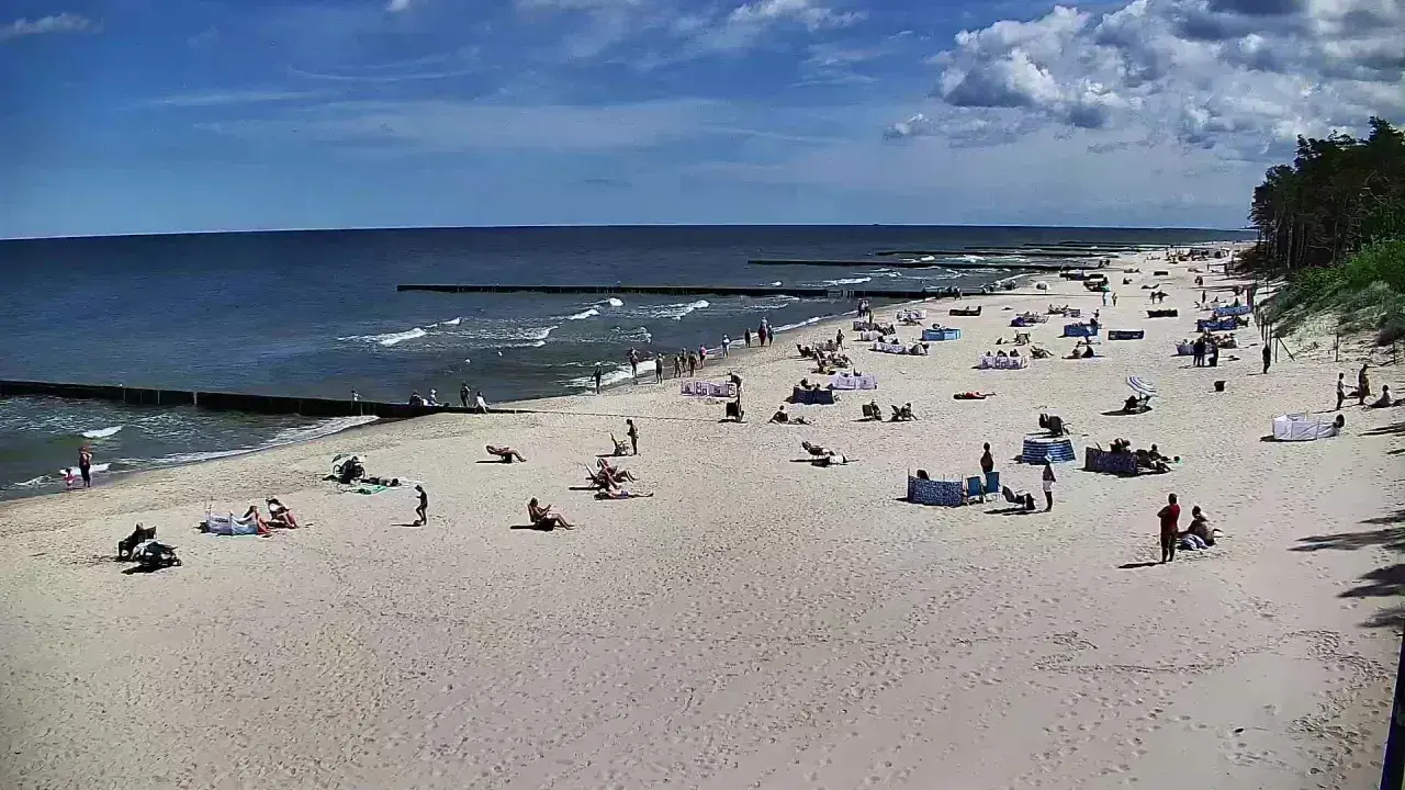 Słoneczny dzień na plaży w Dźwirzynie. Ludzie odpoczywają na piasku, kąpią się w morzu i spacerują po deptaku.