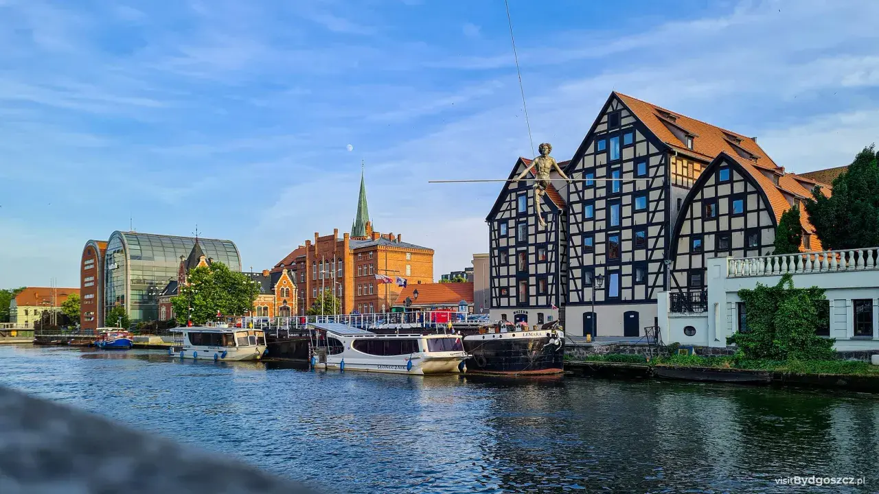 Bydgoszcz wydarzenia dzisiaj: na Wyspie Młyńskiej spaceruje po linie figura, cumują statki, a w tle widać zabytkowe kamienice i nowoczesną architekturę.