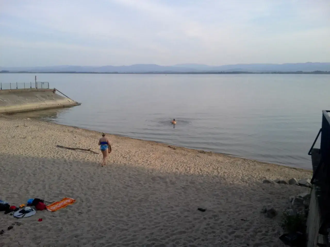 Spokojny dzień nad otmuchowskim jeziorem. Kobieta wchodzi do wody, podczas gdy inna osoba pływa w oddali. Piasek plaży i łagodne fale tworzą relaksującą atmosferę.