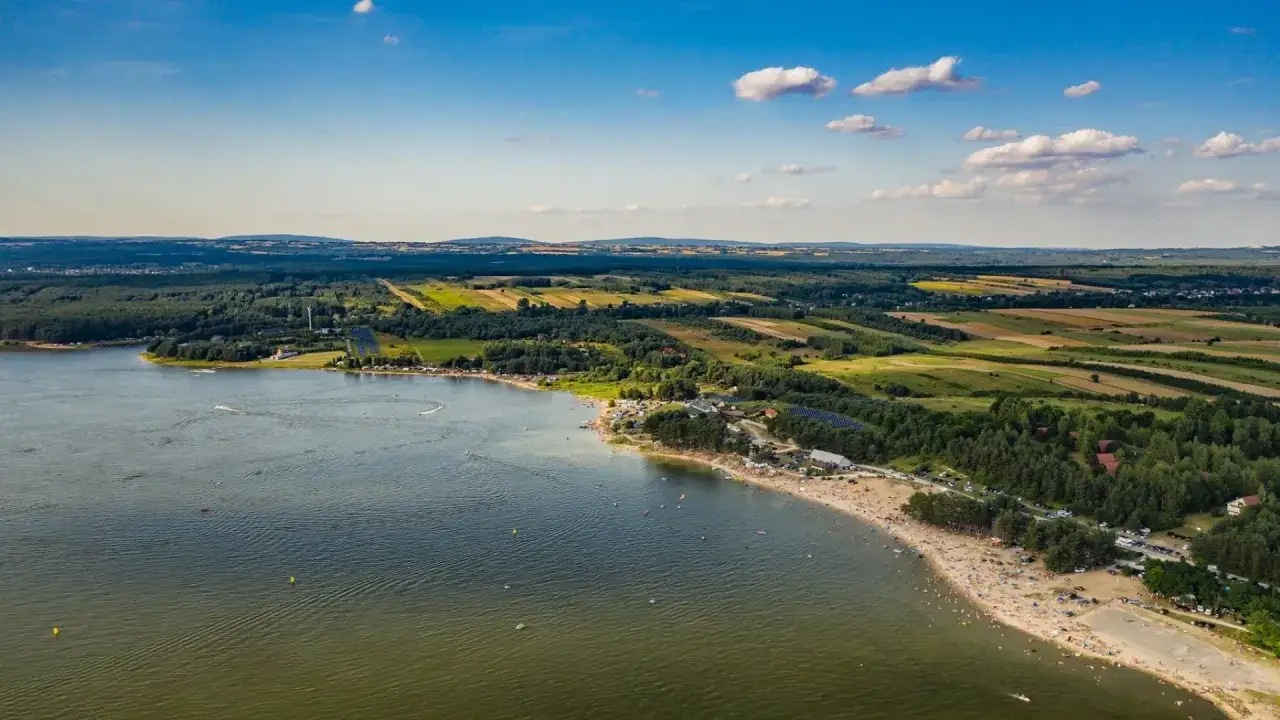 Letnie słońce nad jeziorem Chańcza, pełnym plażowiczów i wodnych atrakcji. W tle zielone lasy i pola uprawne.