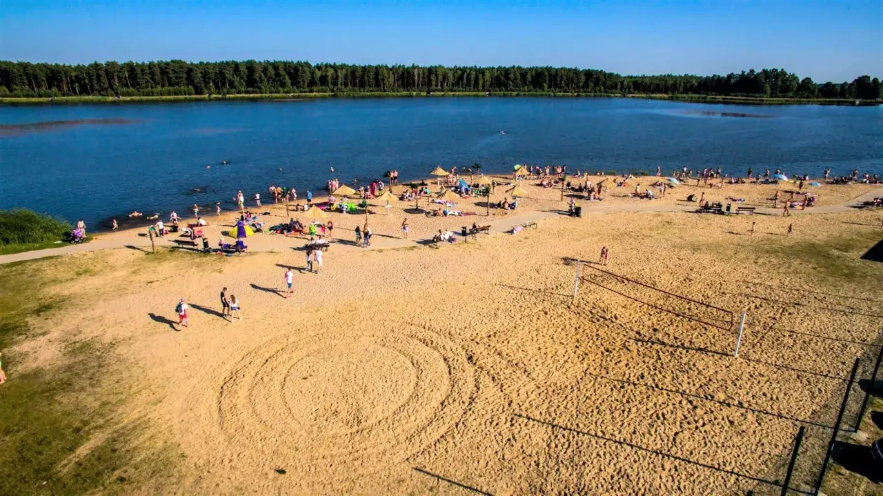 Plaża nad jeziorem, pełna ludzi opalających się i kąpiących. W oddali las, a na piasku boisko do siatkówki. To idealne miejsce na letni wykrot.
