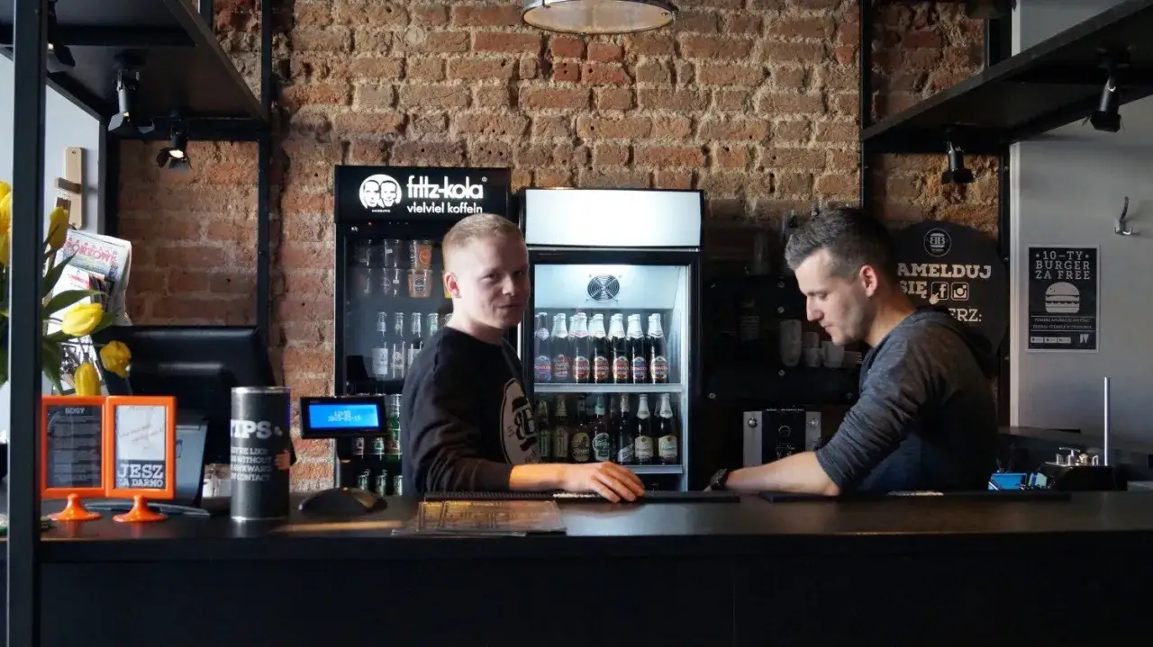 Pracownicy w Bobby Burger Bydgoszcz za ladą, obok lodówki z napojami i ceglanej ściany.