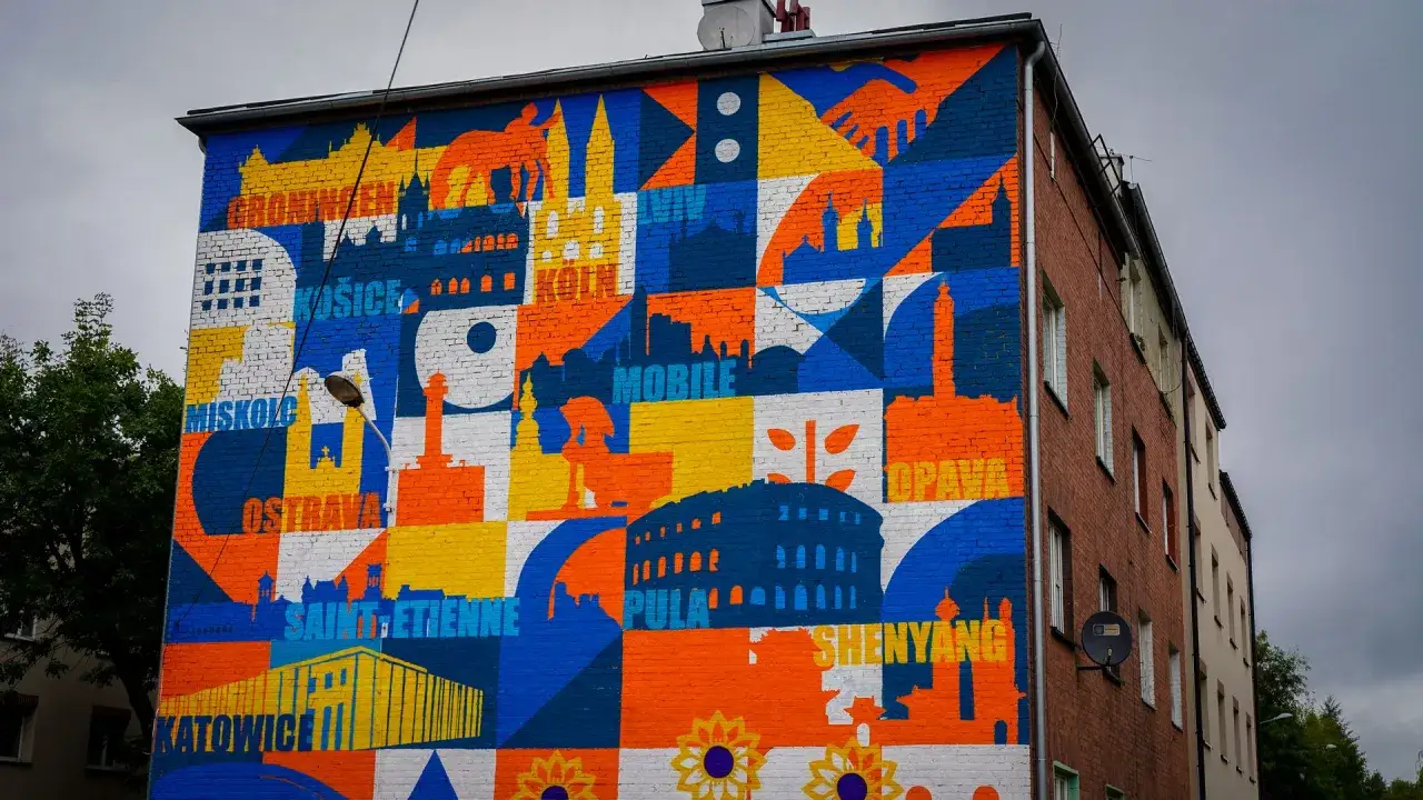 Kolorowy mural na ścianie kamienicy przedstawia sylwetki miast i budynków, w tym Katowice. Nietypowe atrakcje Katowic i innych miast świata na ceglanej ścianie.