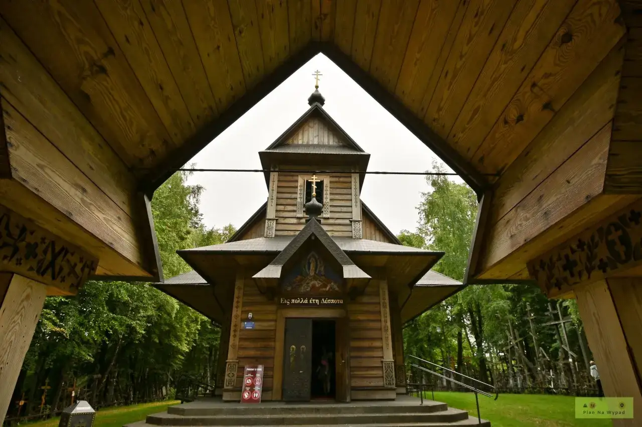 Drewniana cerkiew z krzyżami na tle zieleni. W oddali widać wiele krzyży, tworzących niezwykłą grabarkę.