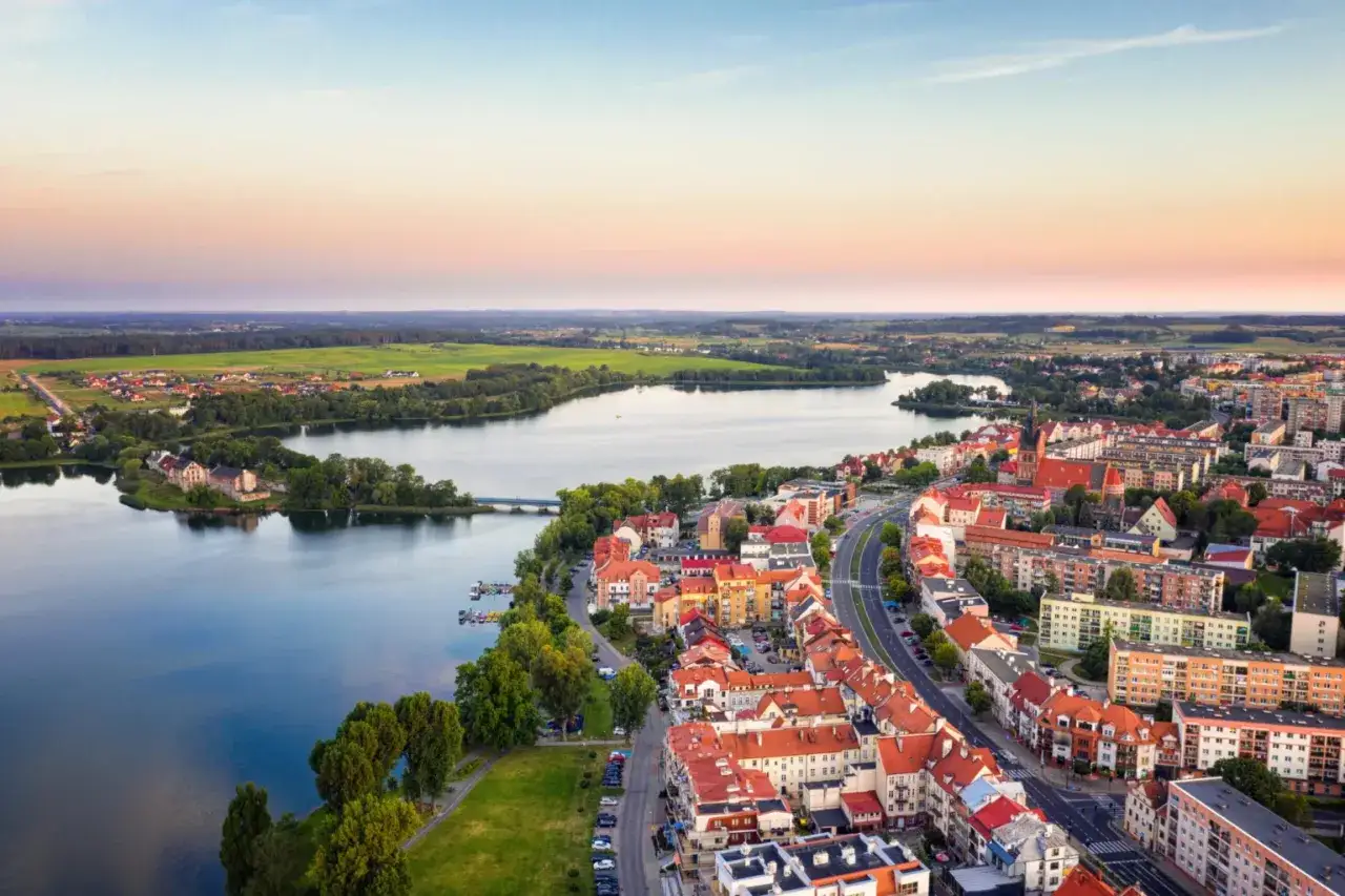 Malownicze miejscowości na Mazurach nad jeziorem, z zabudową miejską i zielonymi terenami w tle.