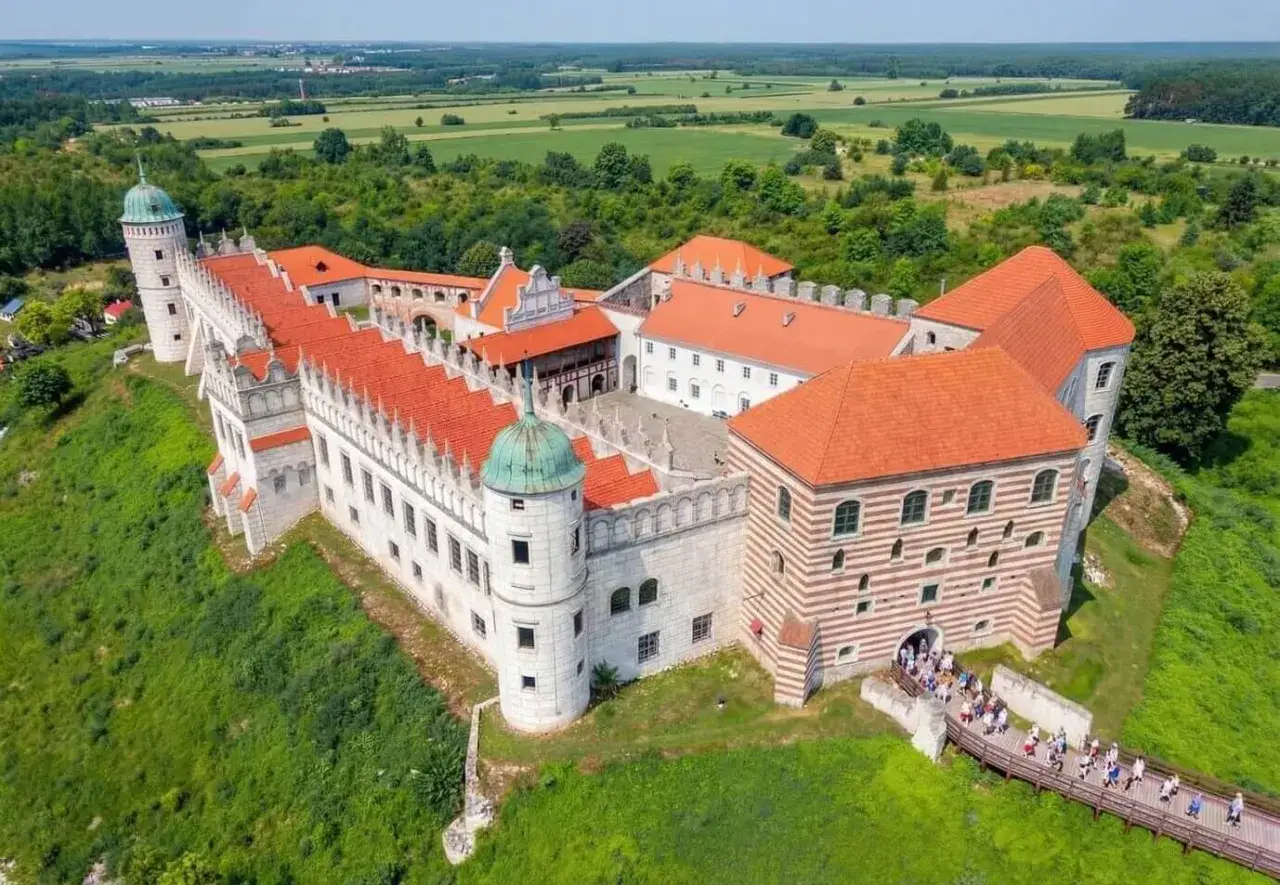 Zamek w Janowcu nad Wisłą, malowniczo położony na wzgórzu, z czerwonymi dachami i zielonymi kopułami, otoczony bujną zielenią.