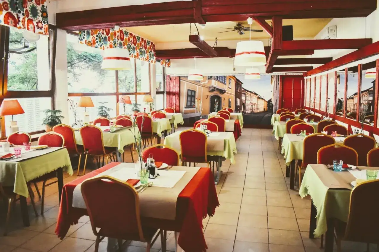 Elegancka sala restauracyjna z czerwonymi krzesłami i stołami nakrytymi na przyjęcie. W tle mural przedstawiający ulicę, jakby z Gdańska.