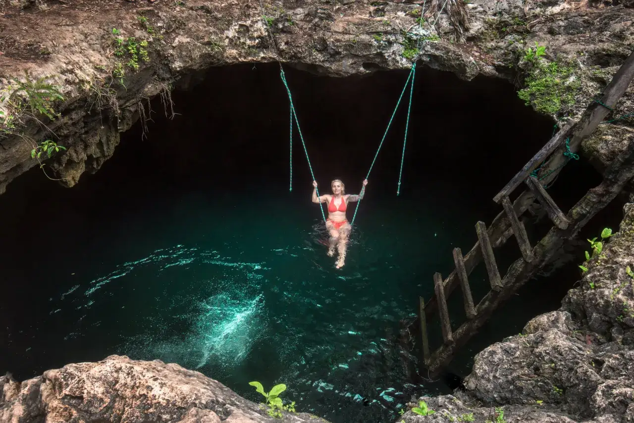 Kobieta na huśtawce w turkusowej wodzie naturalnego basenu – cenote. Drewniana drabinka prowadzi do mrocznej jaskini.