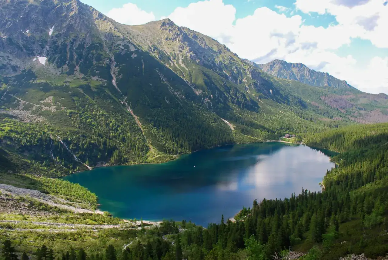 Widok na Morskie Oko z kamery na żywo. Jezioro otoczone górami i lasem, z wodospadem po lewej.
