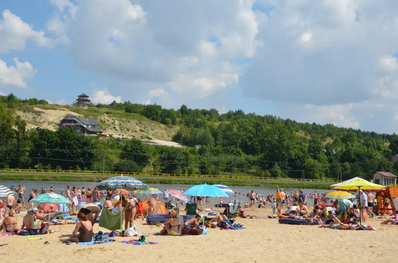 Słoneczny dzień na Krasnobrodzie zalew. Ludzie odpoczywają na plaży, kąpią się w wodzie, pod kolorowymi parasolami.