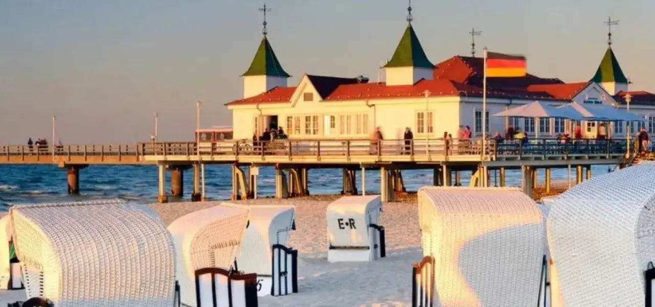 Zachód słońca nad molo w Ahlbecku. Białe kosze plażowe na piasku, a w tle budynek z restauracją i niemiecką flagą. To jedne z wielu atrakcji Ahlbecku.